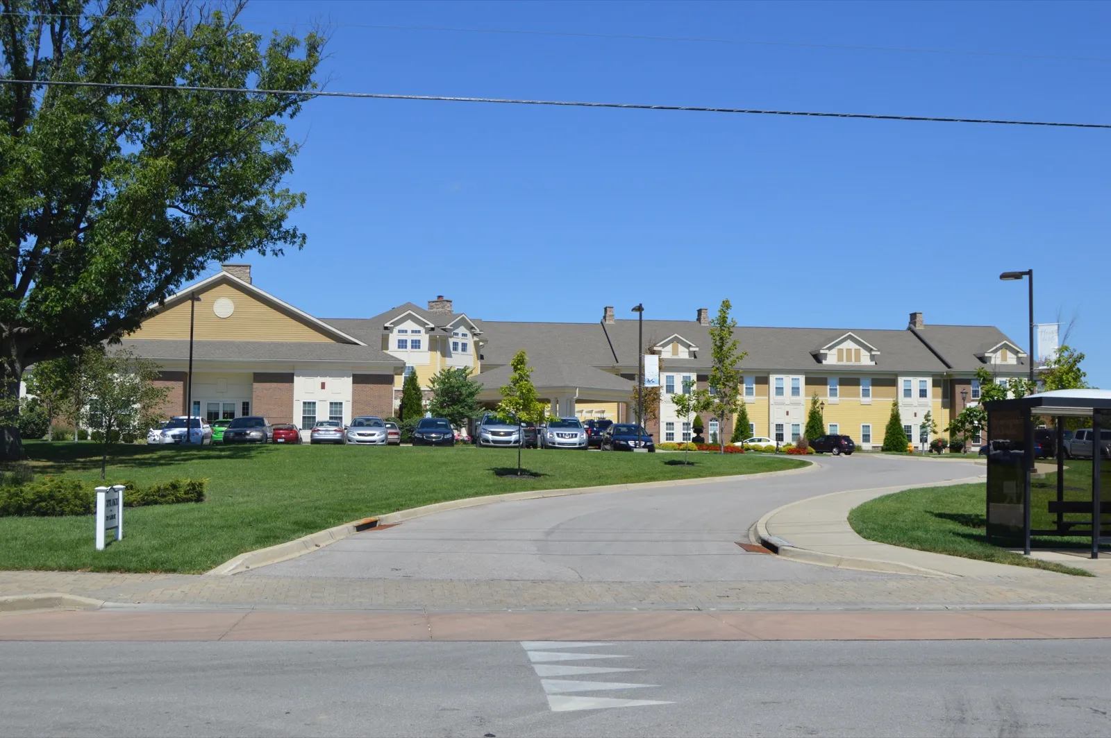 The curving entrance drive of a two-story nursing home with a parking lot in front, under a clear blue sky