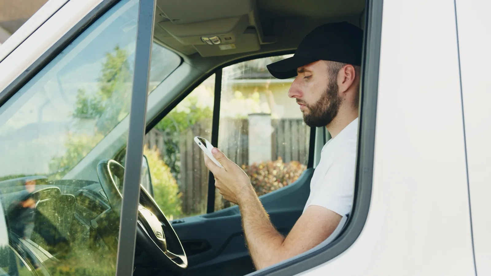 A driver sitting in a parked white van outside a house, looking at his phone while he waits