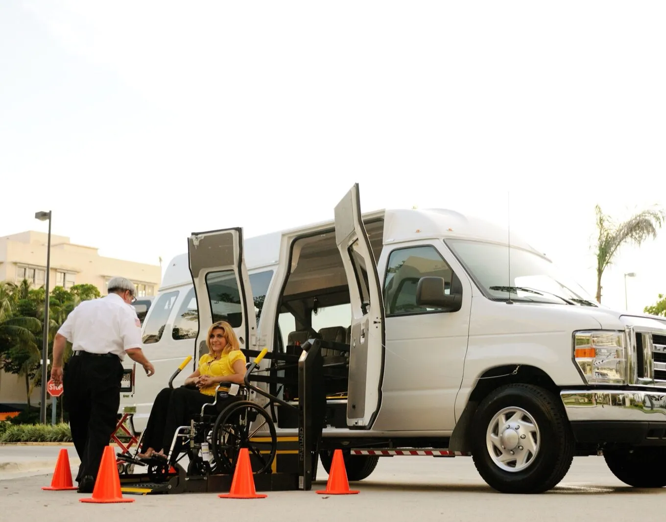 A woman in a wheelchair on the lowered lift of a white high-roof van, with a uniformed driver beside her and orange cones around the lift