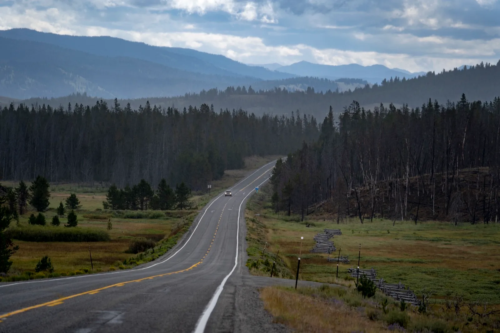 Idaho State Highway 21 curving through a green meadow and pine forest in the Sawtooth National Forest, with mountains under a cloudy sky