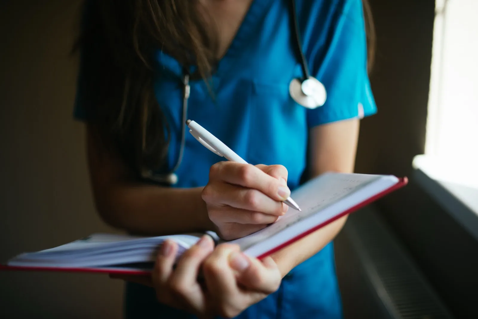 A nurse in blue scrubs with a stethoscope writing in an open notebook with a white pen, face out of frame
