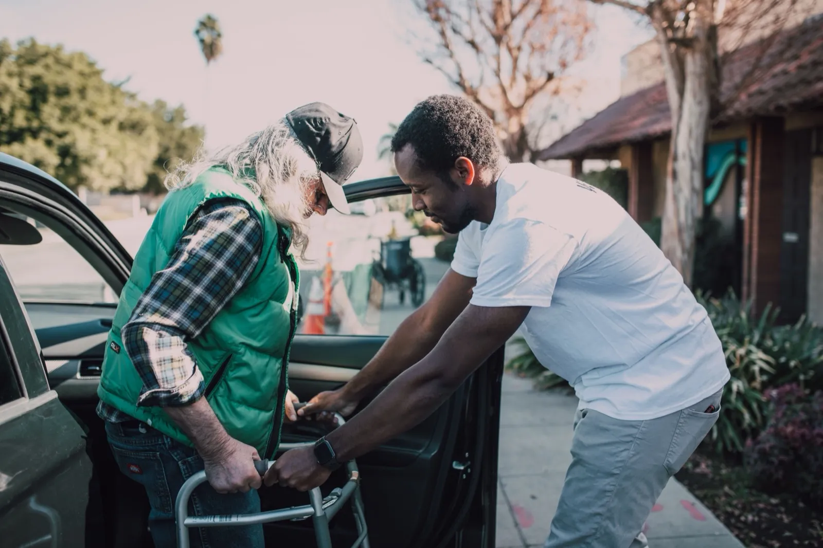 A driver in a white T-shirt steadies a walker as an older man with a cap and green vest steps out of a car at the curb