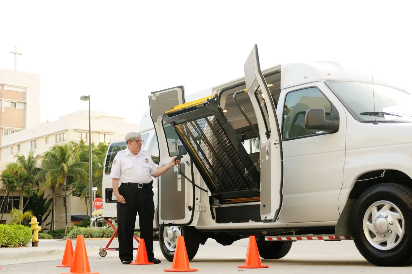 A man in a white uniform shirt operates the raised wheelchair lift of a white van, with orange traffic cones around it