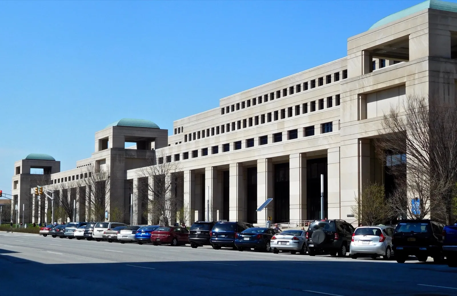 The Indiana Government Center in downtown Indianapolis, a long limestone state office building with green-roofed towers, behind a row of parked cars
