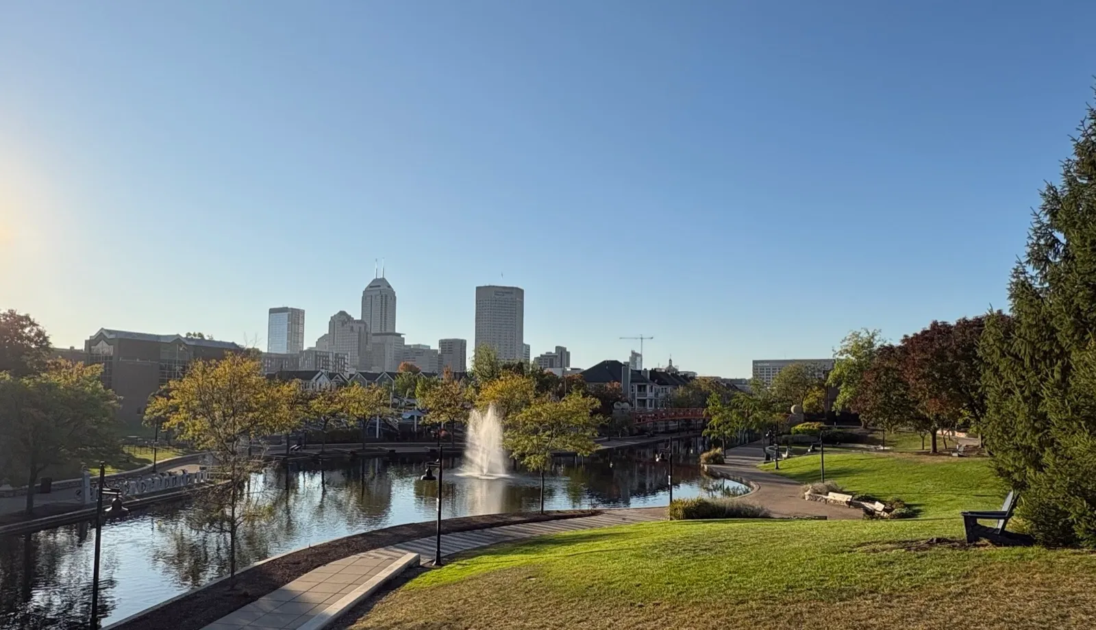 The downtown Indianapolis skyline beyond the Canal Walk, with a fountain in the canal and autumn trees on a clear morning