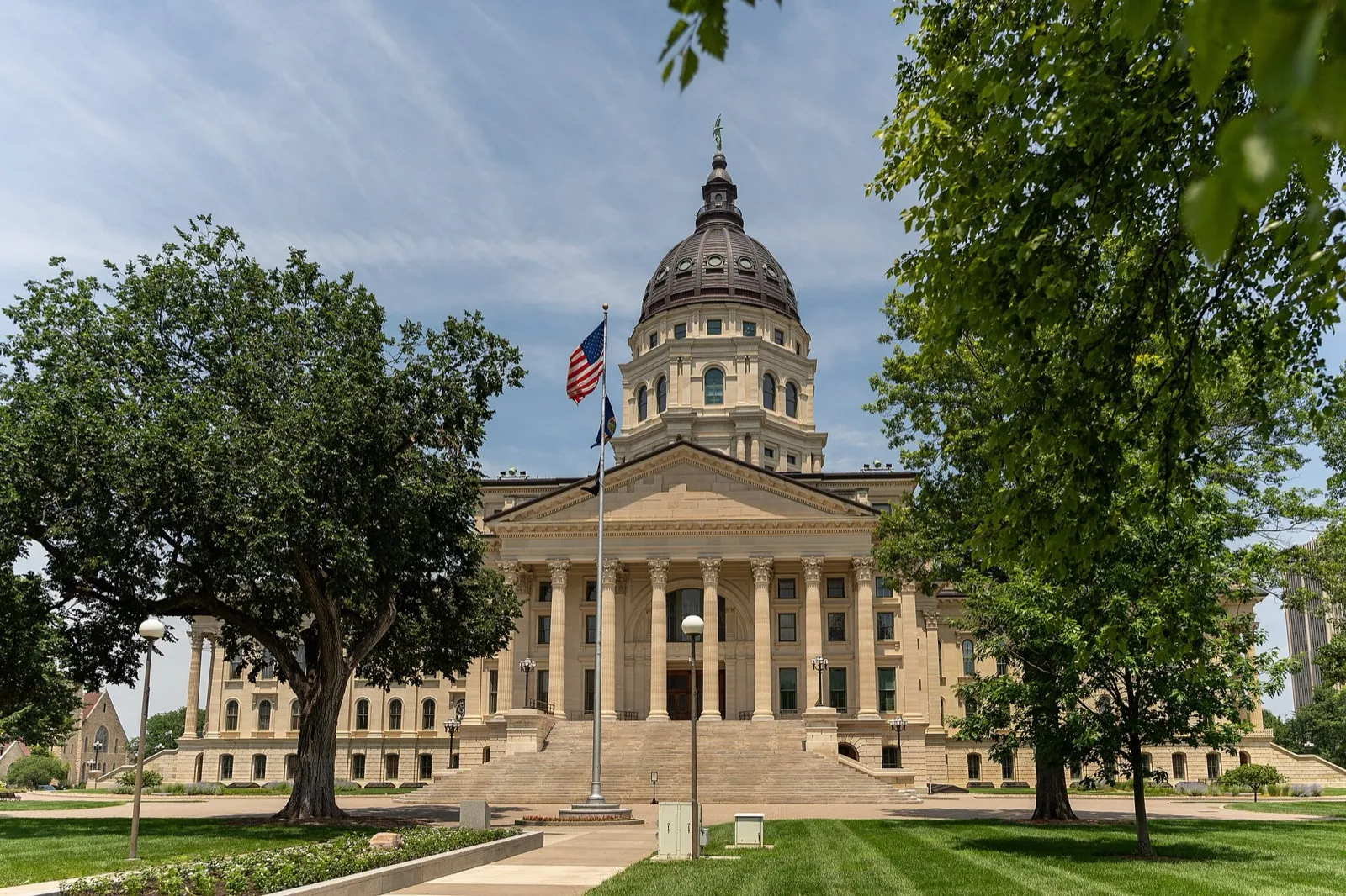 The Kansas State Capitol in Topeka on a sunny summer day, framed by trees, with flags flying on the lawn out front