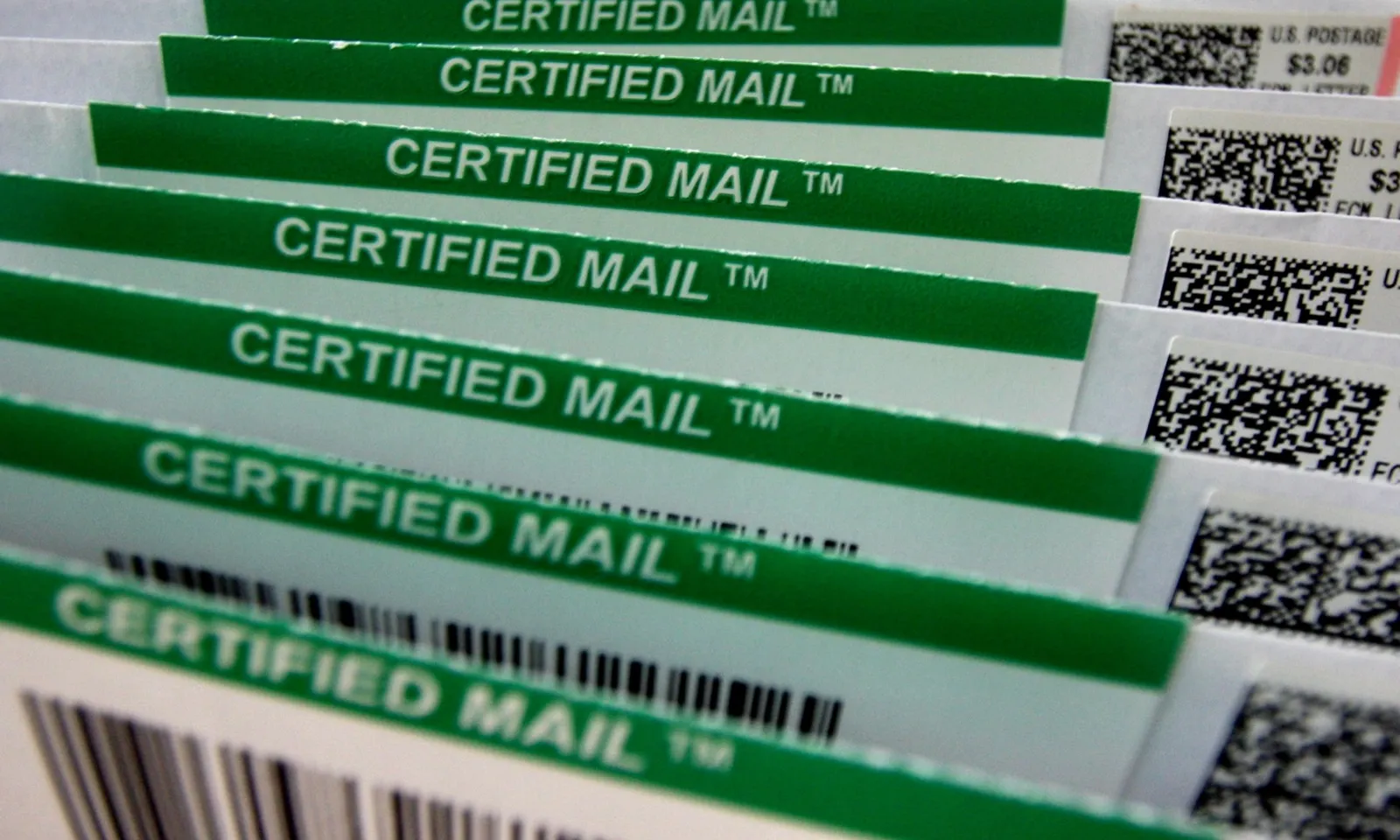 A row of envelopes in a mail tray, each with a green Certified Mail label and a postage strip