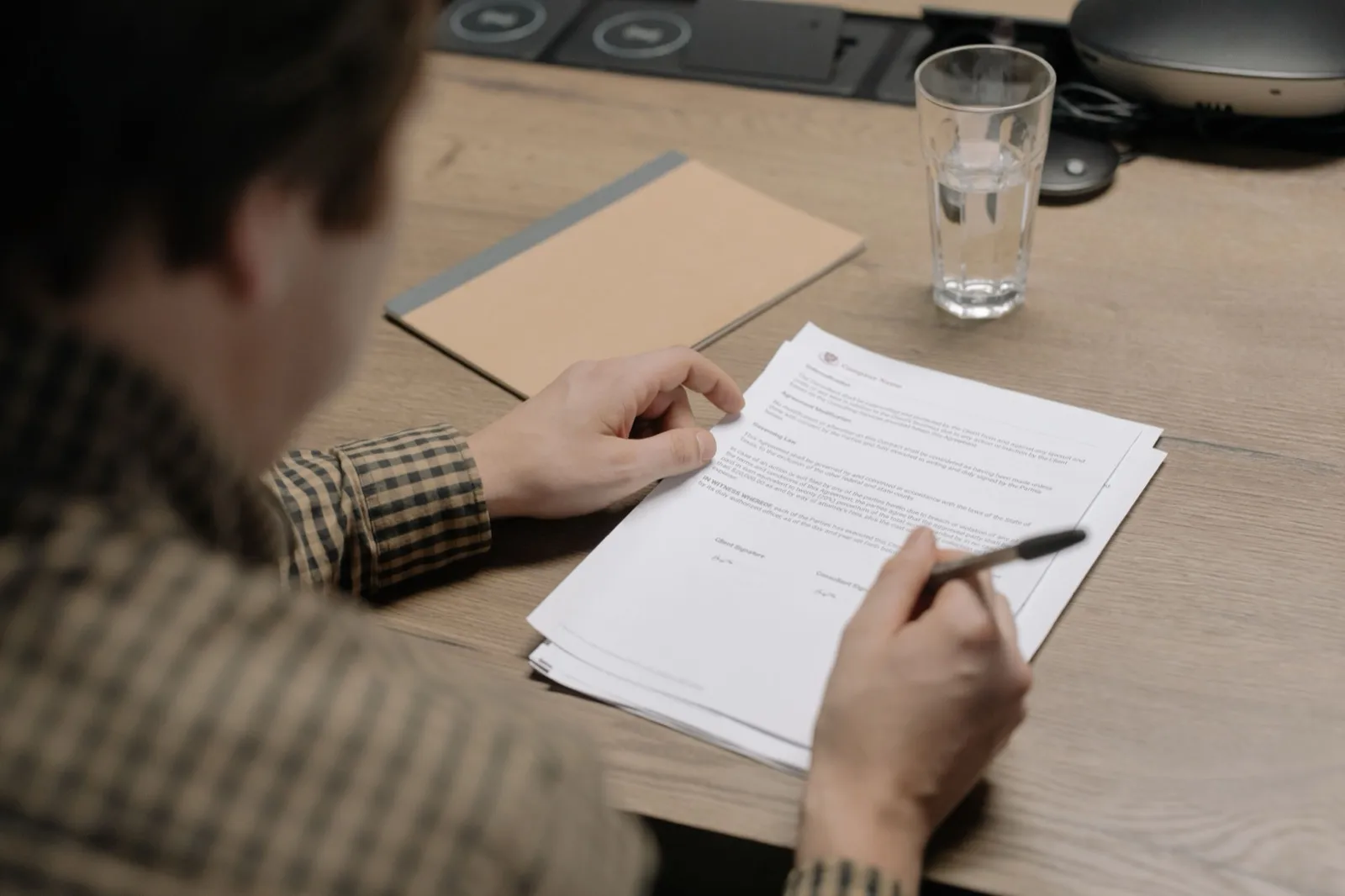 A man at a wooden table holding a pen over a printed contract, with a notebook and a glass of water beside it