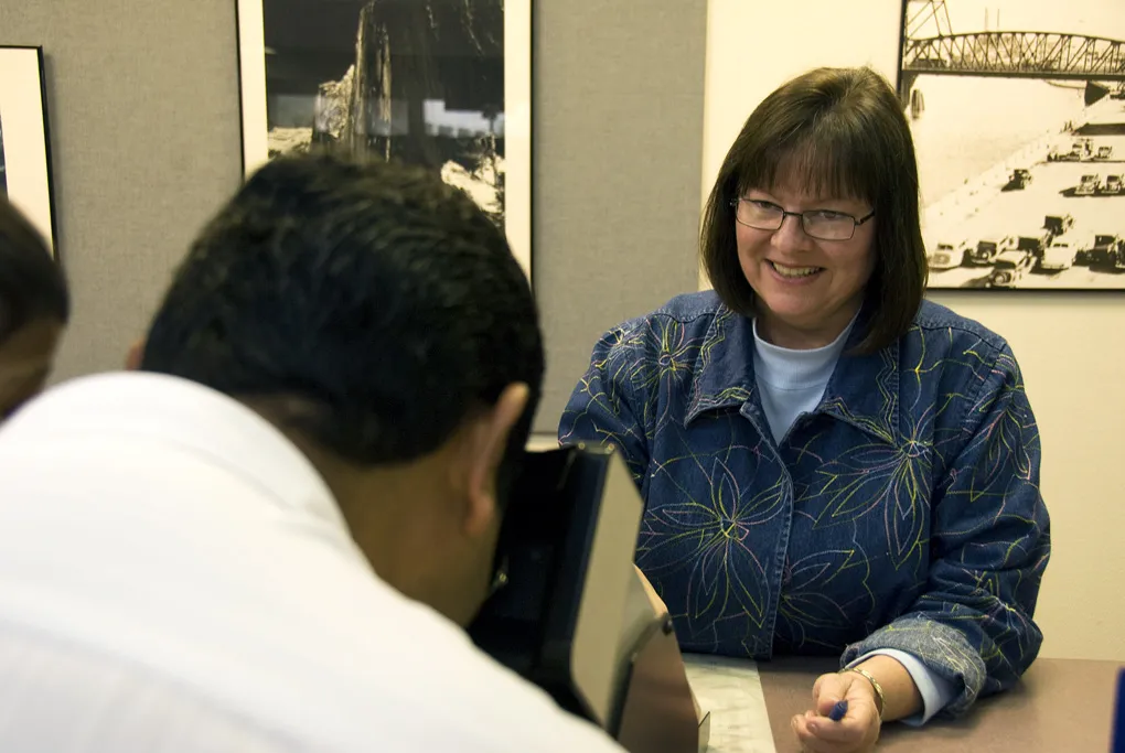 A license applicant looks into a vision testing machine at a licensing office counter while a clerk waits with a pen