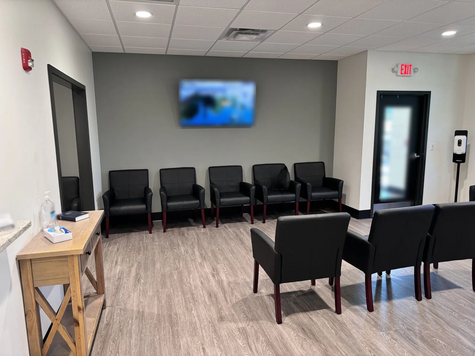 An empty clinic waiting room with rows of dark chairs, a wall television, and a glass exit door