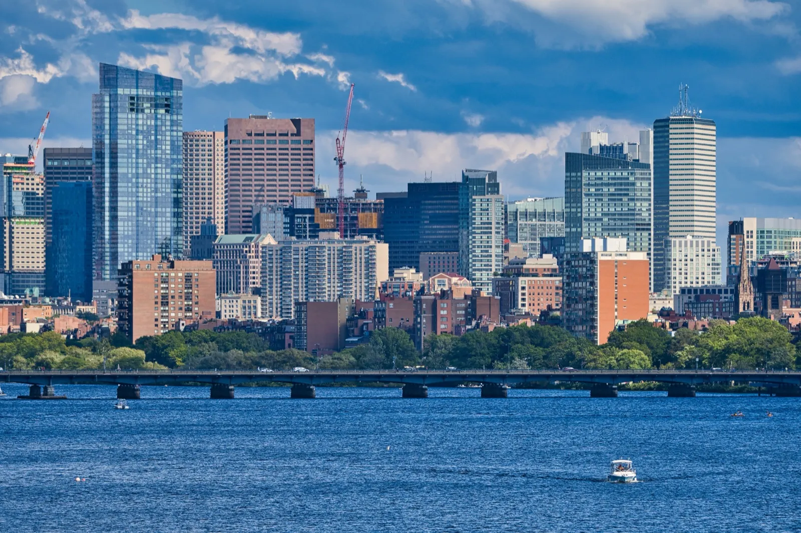 The Boston skyline above the Charles River on a sunny day, with a low bridge across the water and a small boat in the foreground