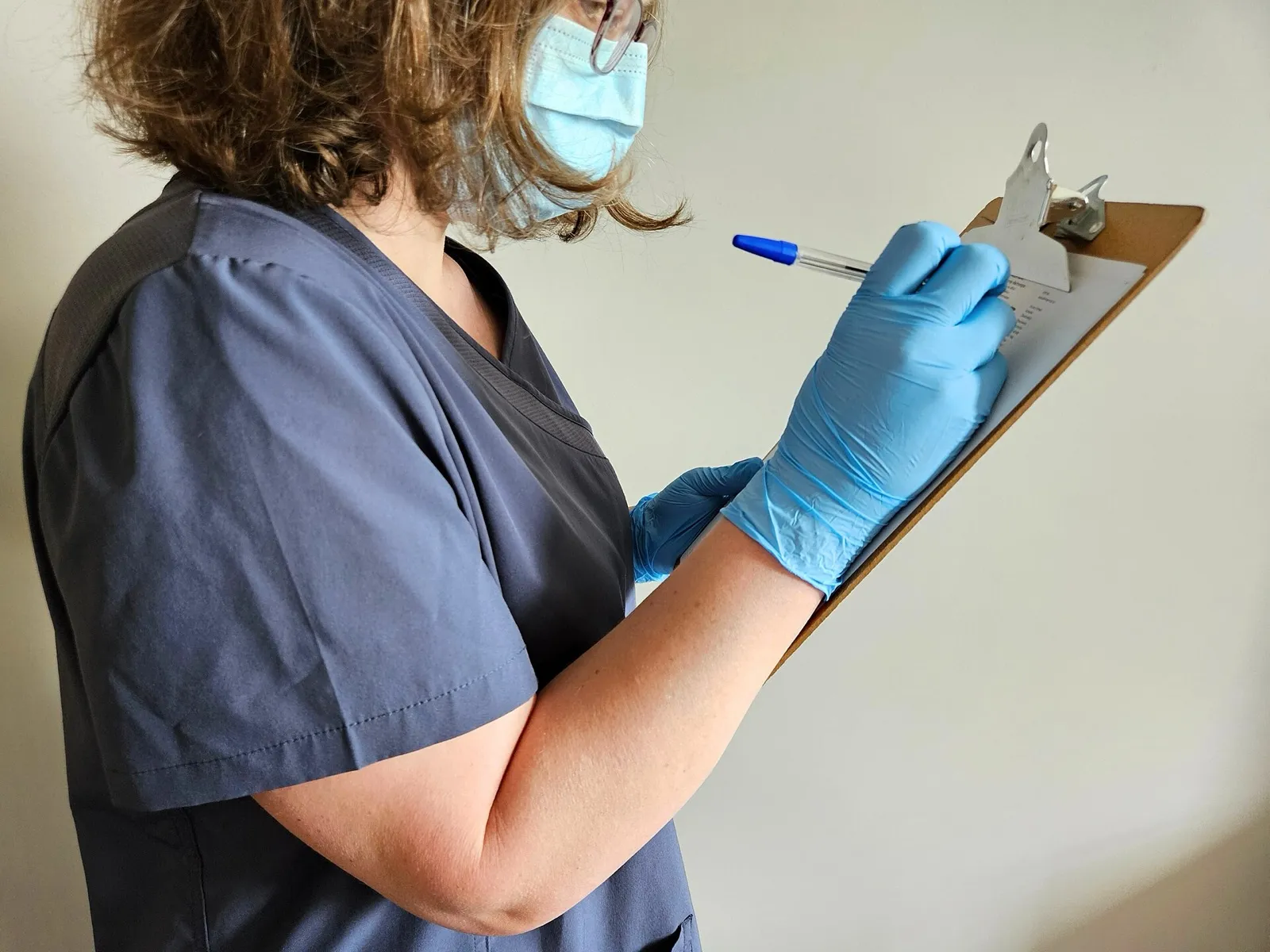 A health care worker in gray scrubs, a face mask, and blue gloves filling out a form on a clipboard
