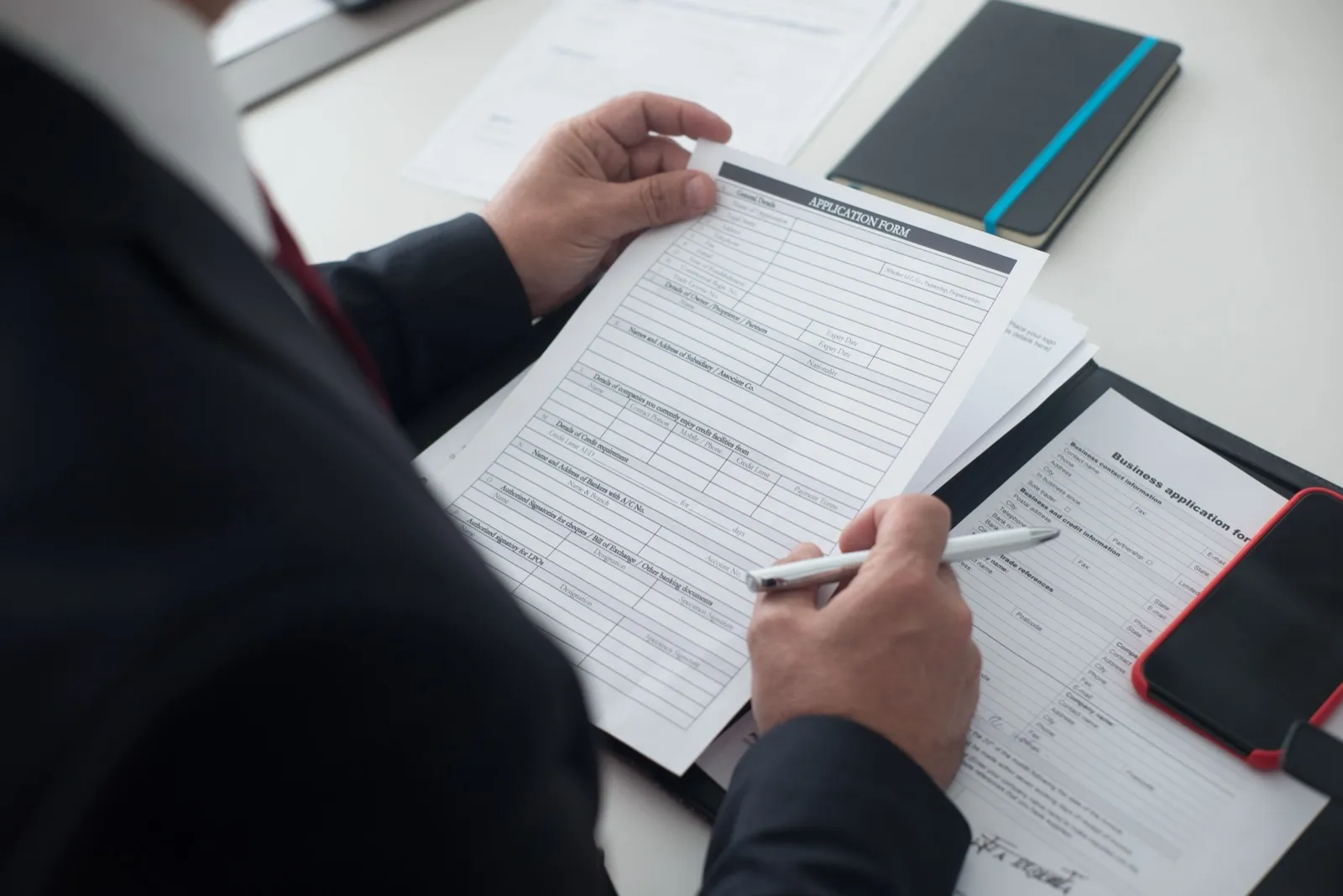 A person in a dark suit at a desk reviews a printed application form with a pen, with a second business application form beside it