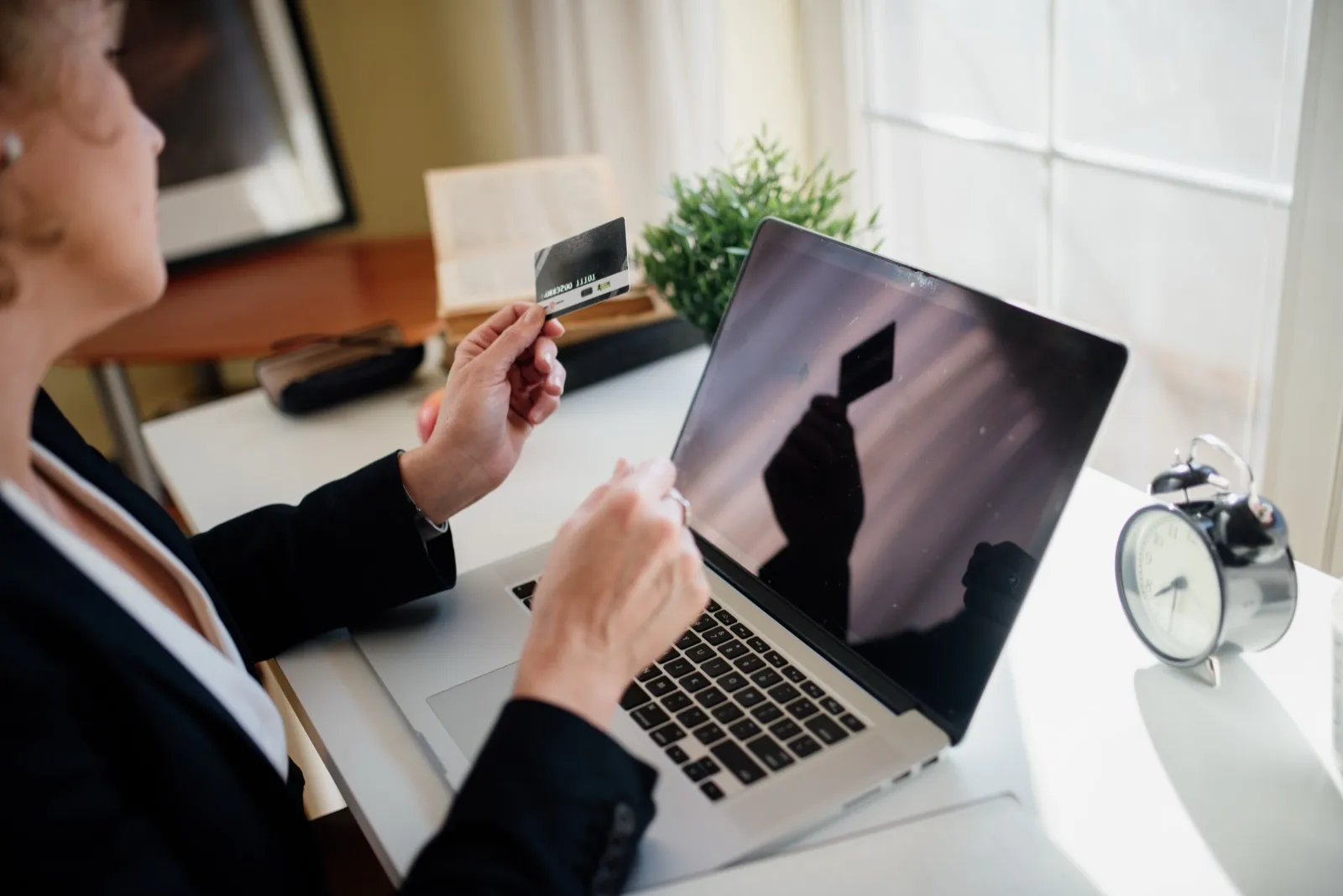 A woman at a white desk holds a payment card in front of an open laptop, with an alarm clock beside it