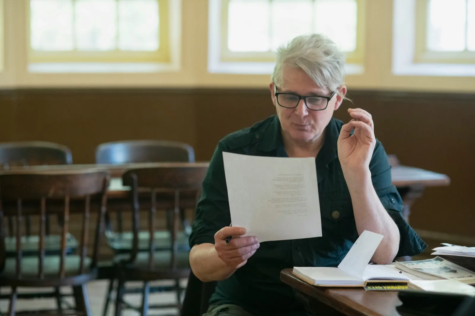 A man with gray hair and glasses sits at a wooden table and reads a printed letter