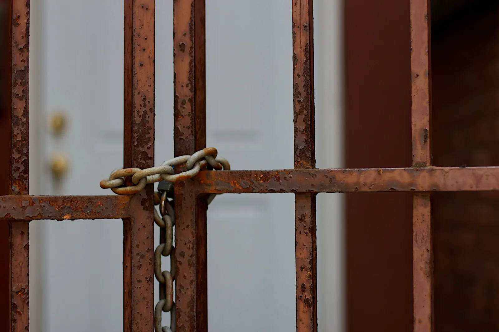 A rusted iron gate closed with a chain and padlock, with a white door blurred behind the bars