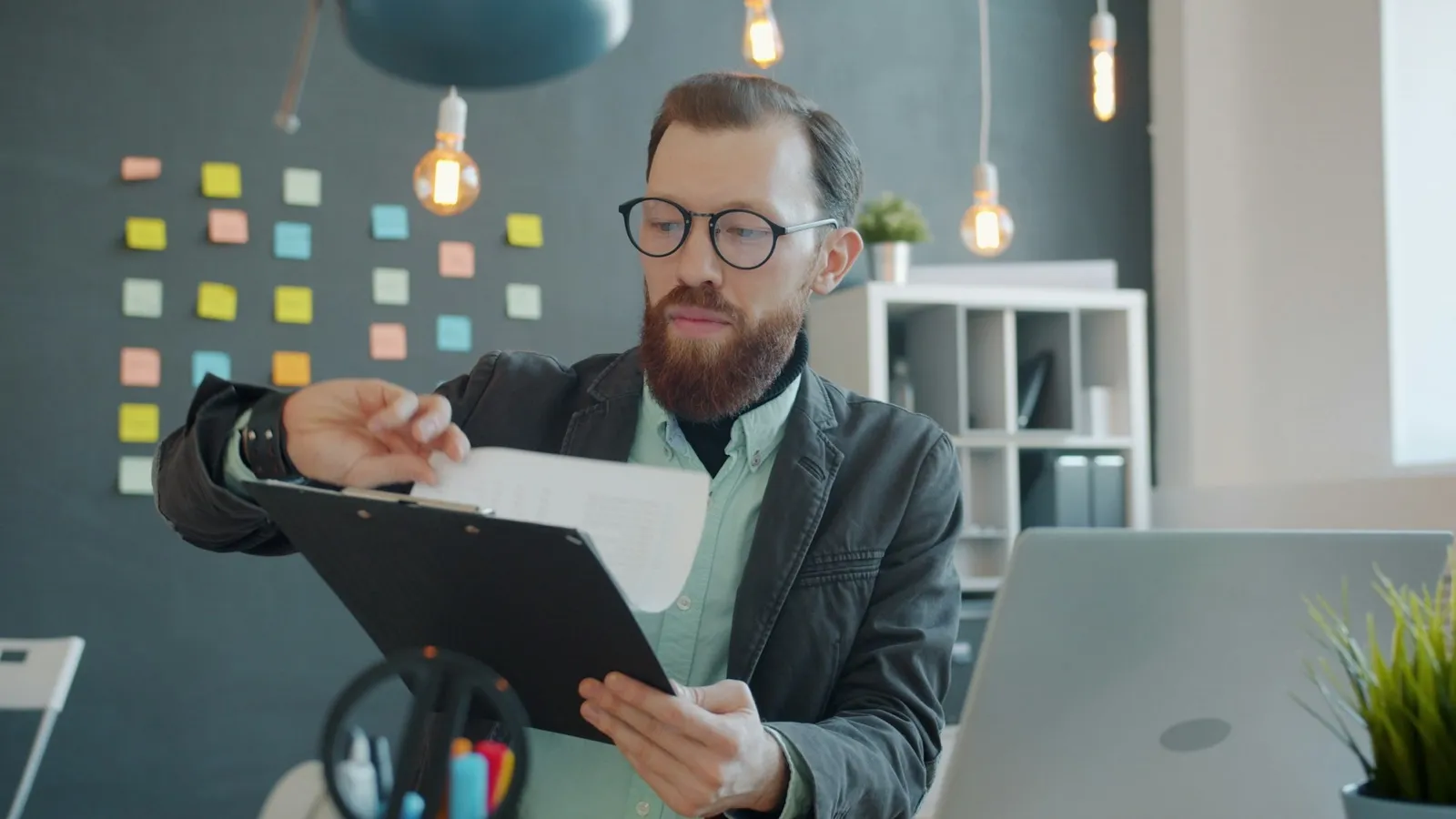 A man in glasses at an office desk reads a printed page on a clipboard, with a laptop open beside him