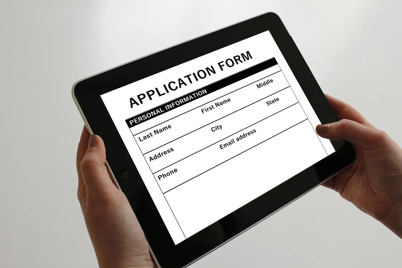 Two hands holding a tablet that shows a blank application form with fields for name, address, and phone