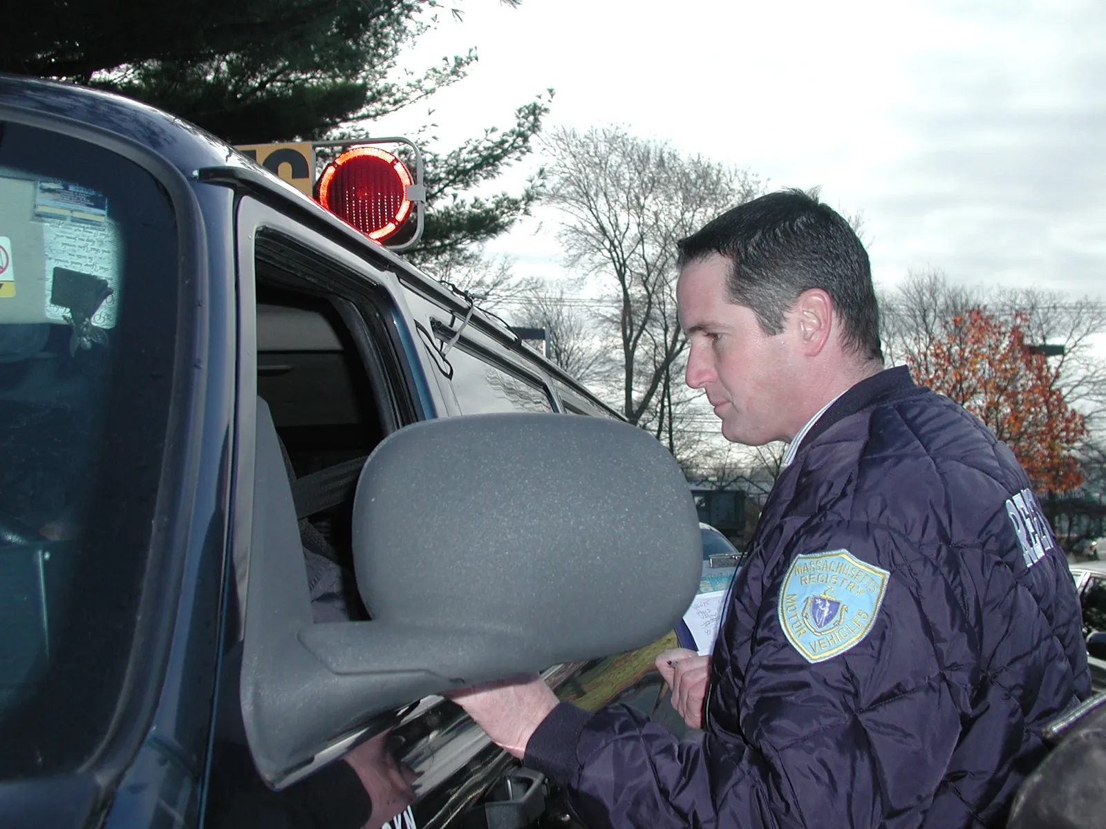 A state motor vehicle inspector with a pen and paperwork checks a school transport van through its open driver window during a safety inspection