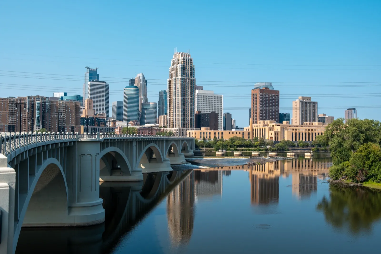 Downtown Minneapolis seen across the Mississippi River, with the arches of the Third Avenue Bridge on the left and the towers reflected in the water