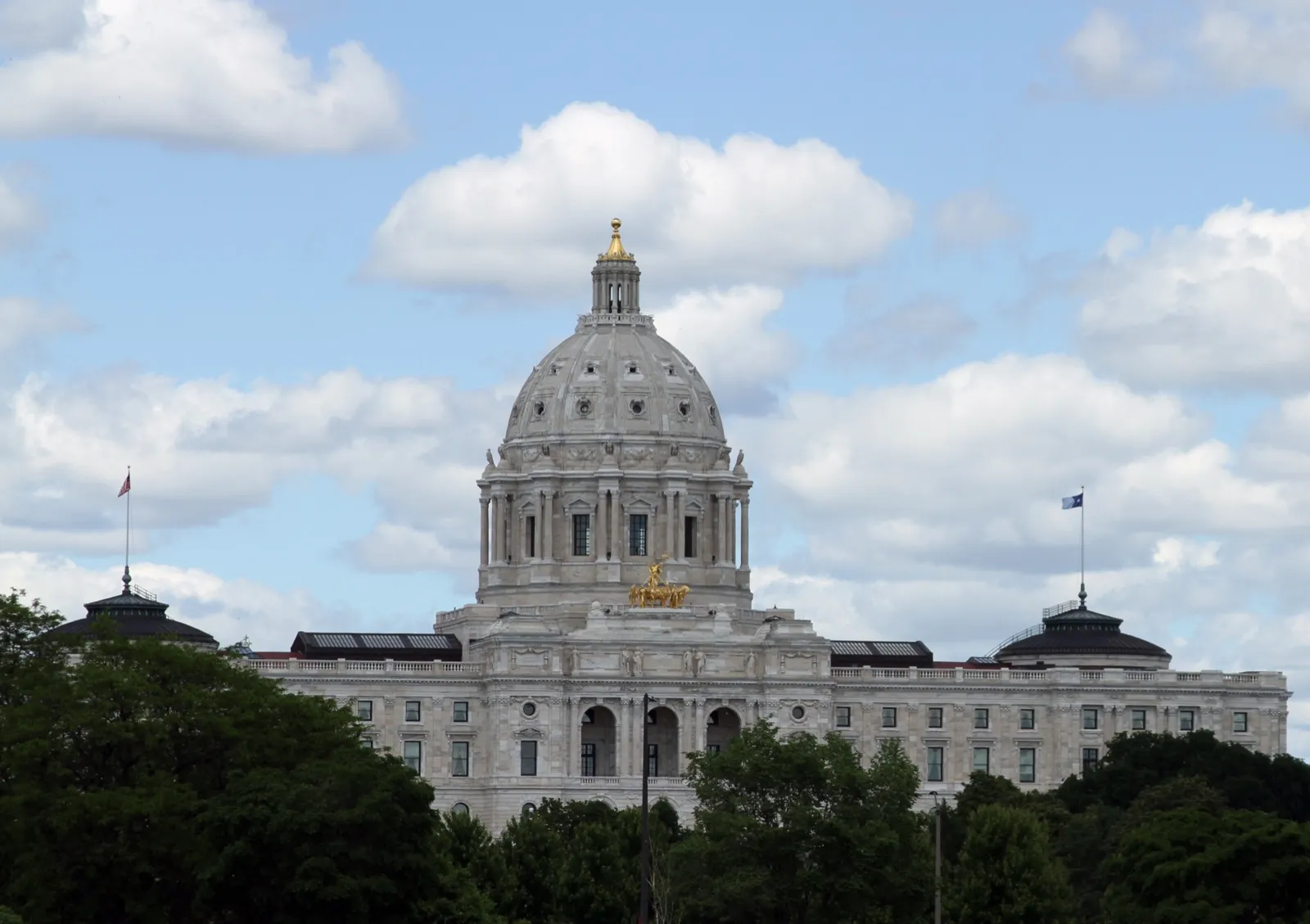 The white marble dome of the Minnesota State Capitol in St. Paul above a line of green trees, under a partly cloudy sky