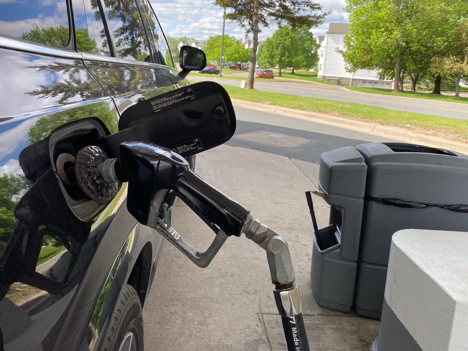 A black fuel nozzle in the open fuel door of a dark car at a gas pump, with trees and a quiet street behind it
