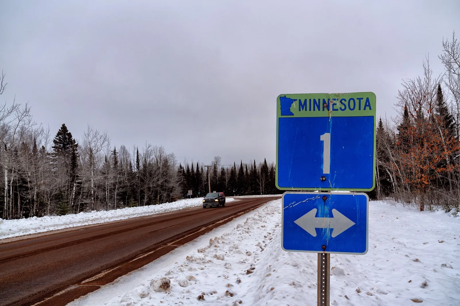 A blue Minnesota Highway 1 route sign beside a snowy two-lane road through pine and birch woods near Silver Bay
