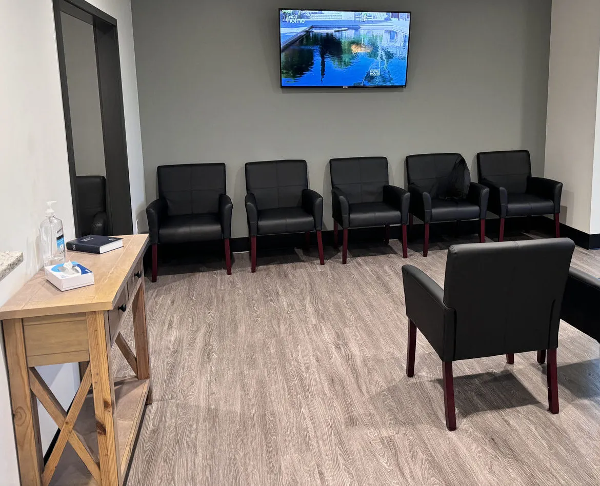 An empty clinic waiting room with rows of dark padded chairs, a wall television, and a wood side table