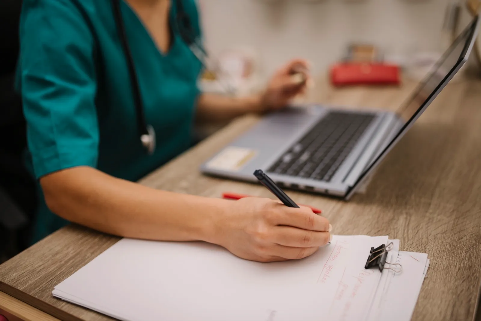A health care worker in teal scrubs with a stethoscope writes on a stack of forms at a desk beside a laptop, face out of frame