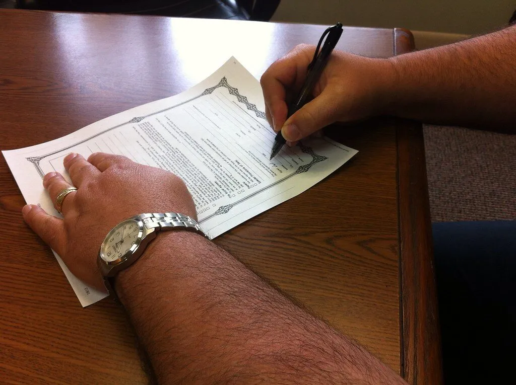 A person at a wooden desk signing a printed form with a black pen