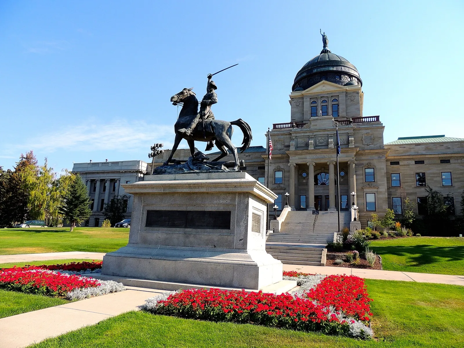 The Montana State Capitol in Helena on a sunny day, with an equestrian statue and red flower beds on the lawn in front