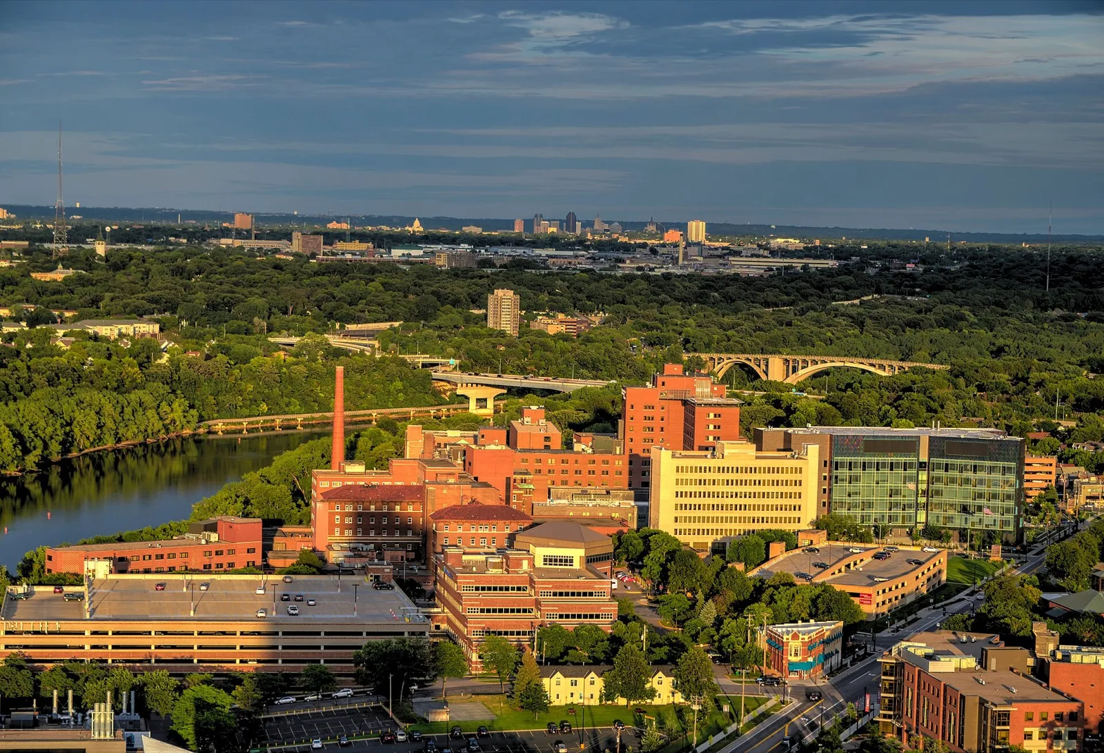 A large brick and glass medical center campus beside a river, seen from above, with a city skyline far in the distance