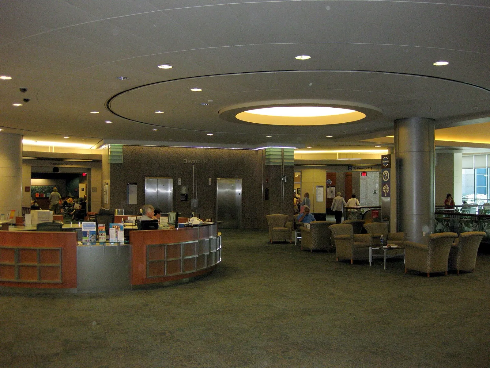 A hospital clinic lobby with staff at a curved reception desk with computers, and patients waiting in armchairs nearby