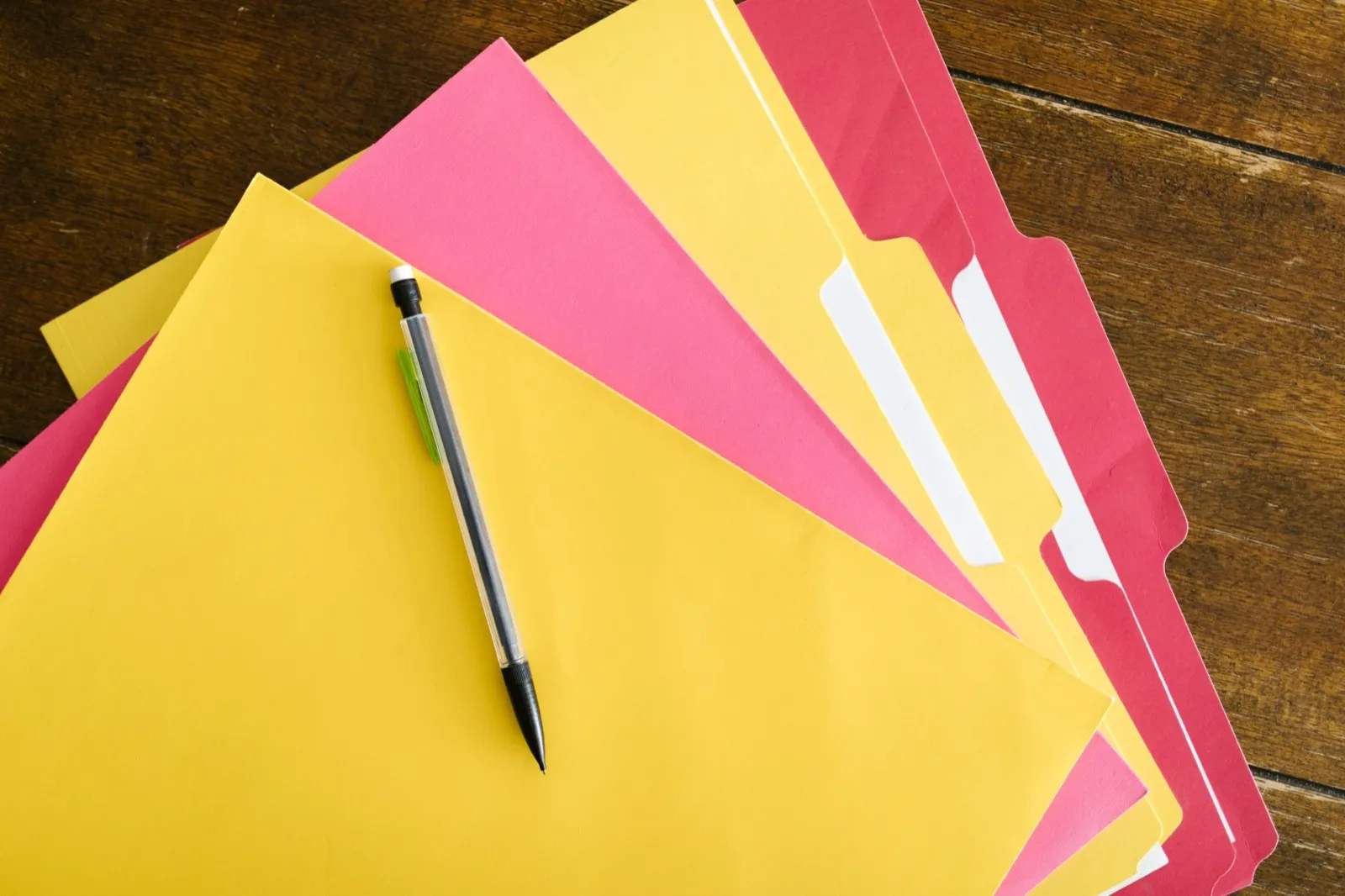 Yellow, pink, and red file folders fanned out on a wooden table with a mechanical pencil on top