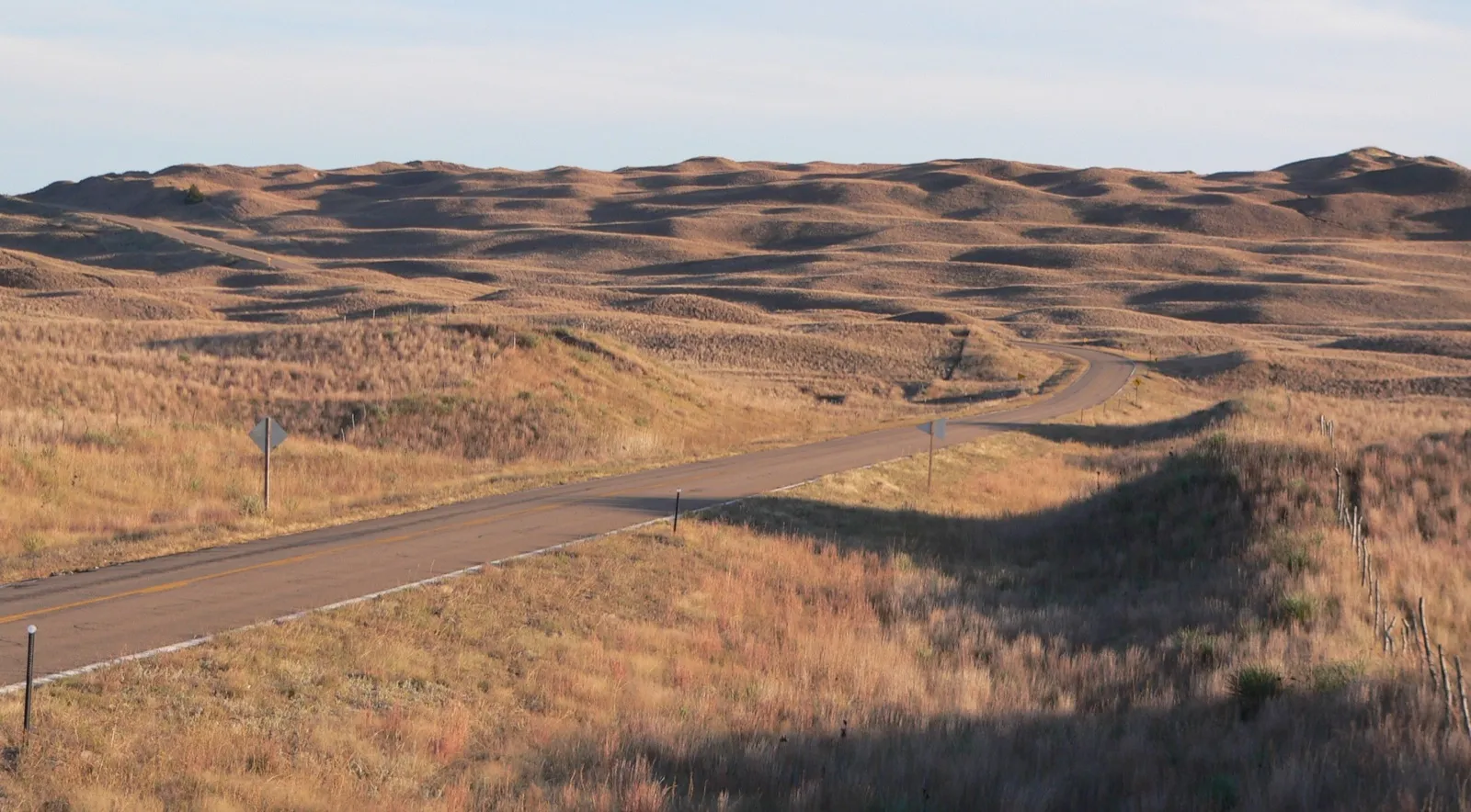A two-lane state highway curving through the grass-covered Sandhills of Hooker County, Nebraska, in late afternoon light
