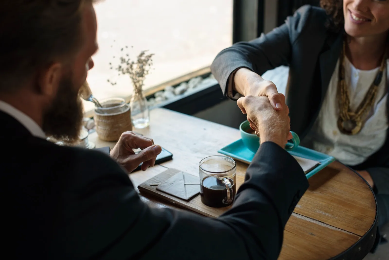 Two people in business clothes shaking hands across a small wooden cafe table with cups of coffee