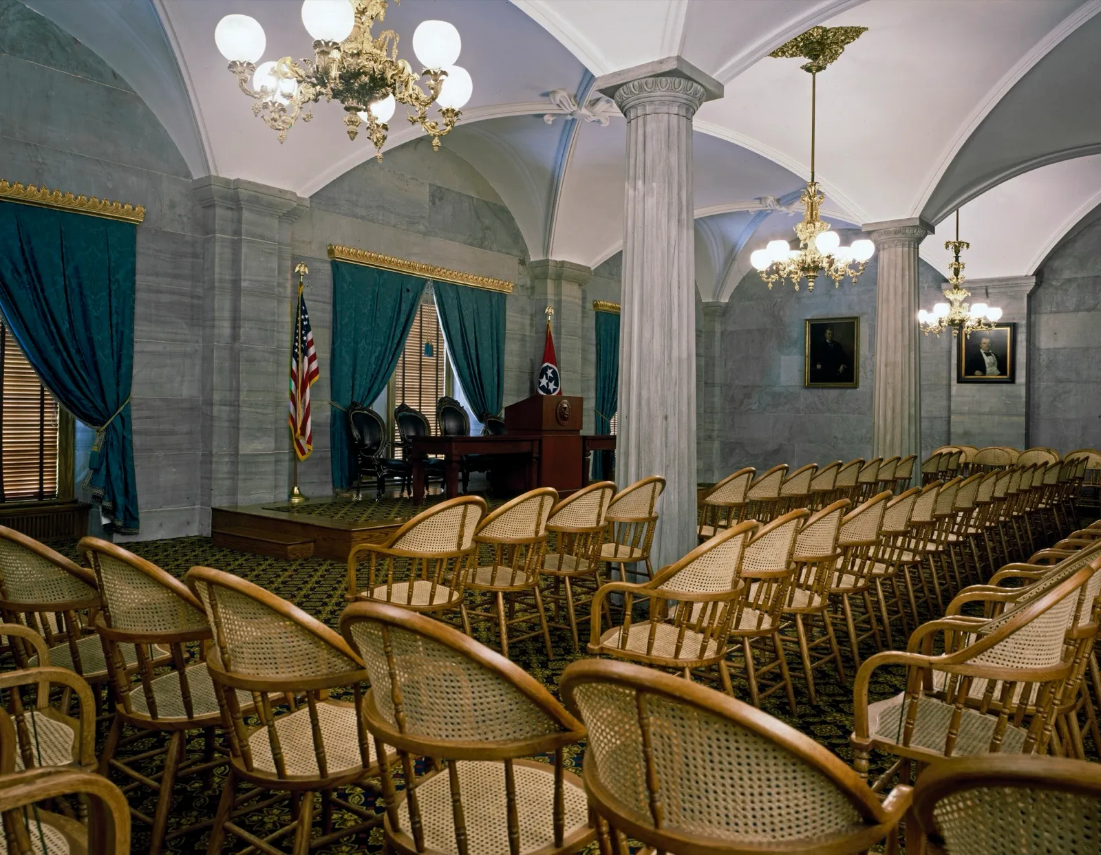 An empty legislative hearing room at the Tennessee Capitol, with rows of cane chairs facing a raised desk, flags, and chandeliers