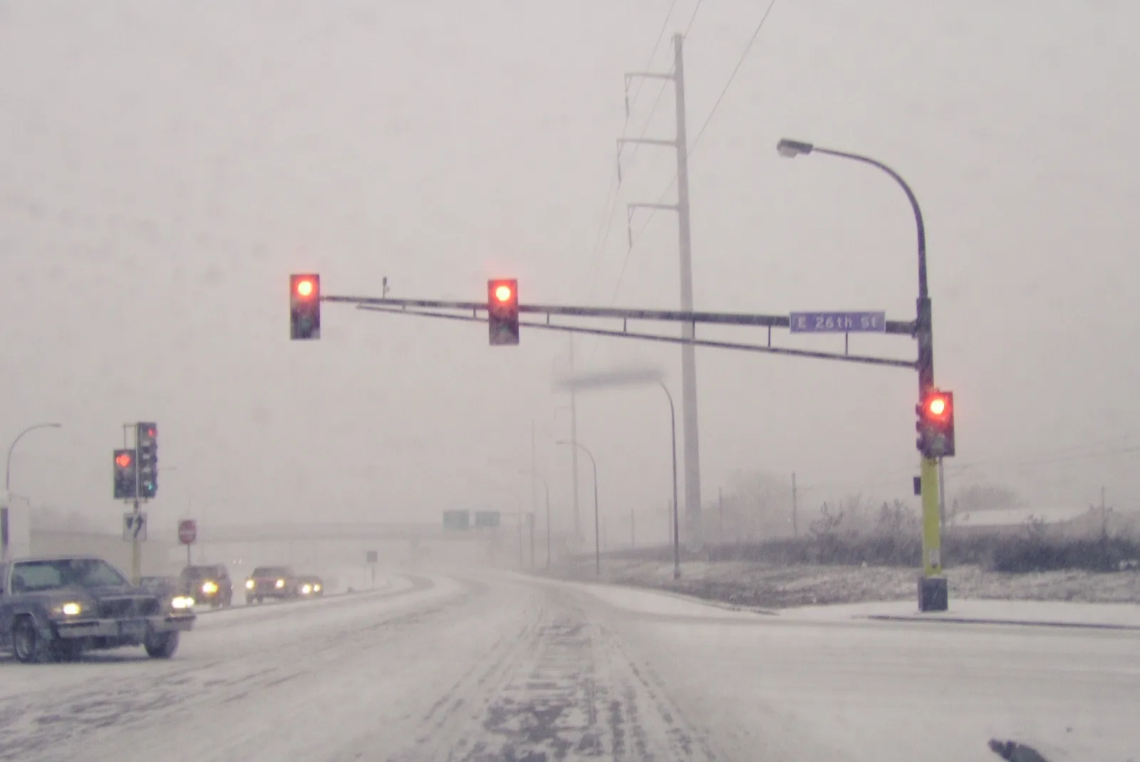 A snow-covered city intersection in a heavy snowstorm, seen through a car windshield, with red traffic lights and oncoming cars using their headlights