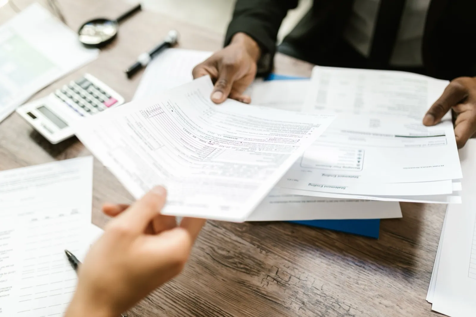 One person handing a printed form across a wooden desk to another, with a calculator and a stack of statements beside them