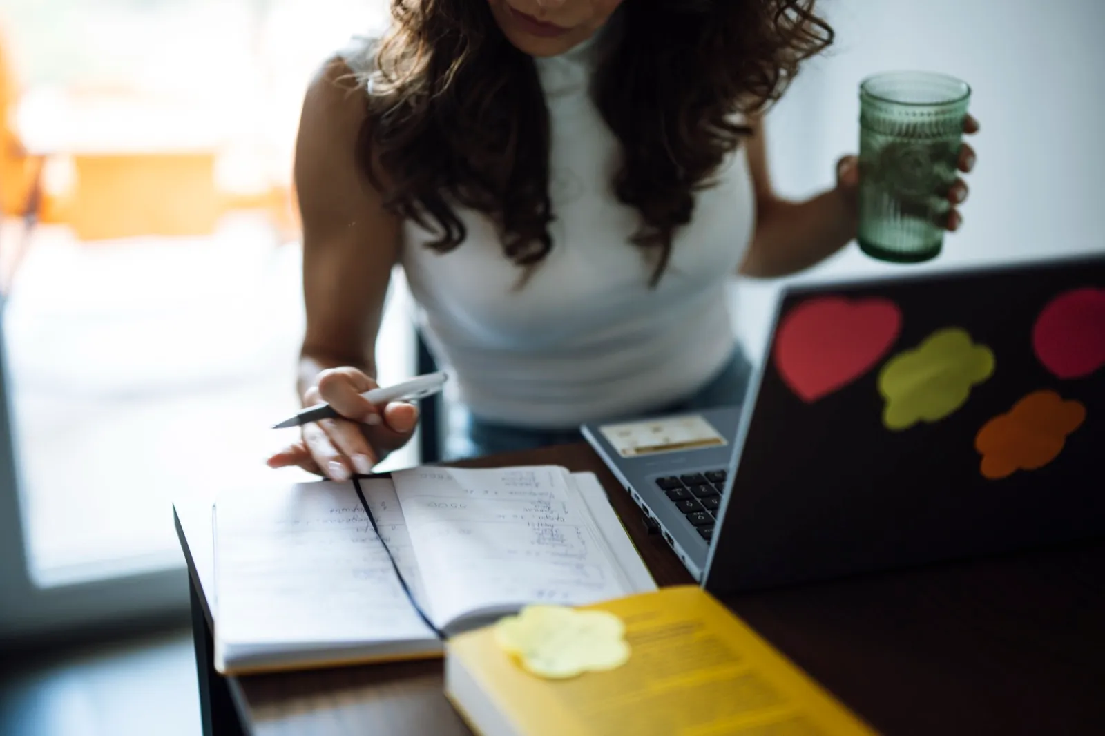 A person at a desk writing notes in an open notebook beside a laptop, with a yellow book in front of them
