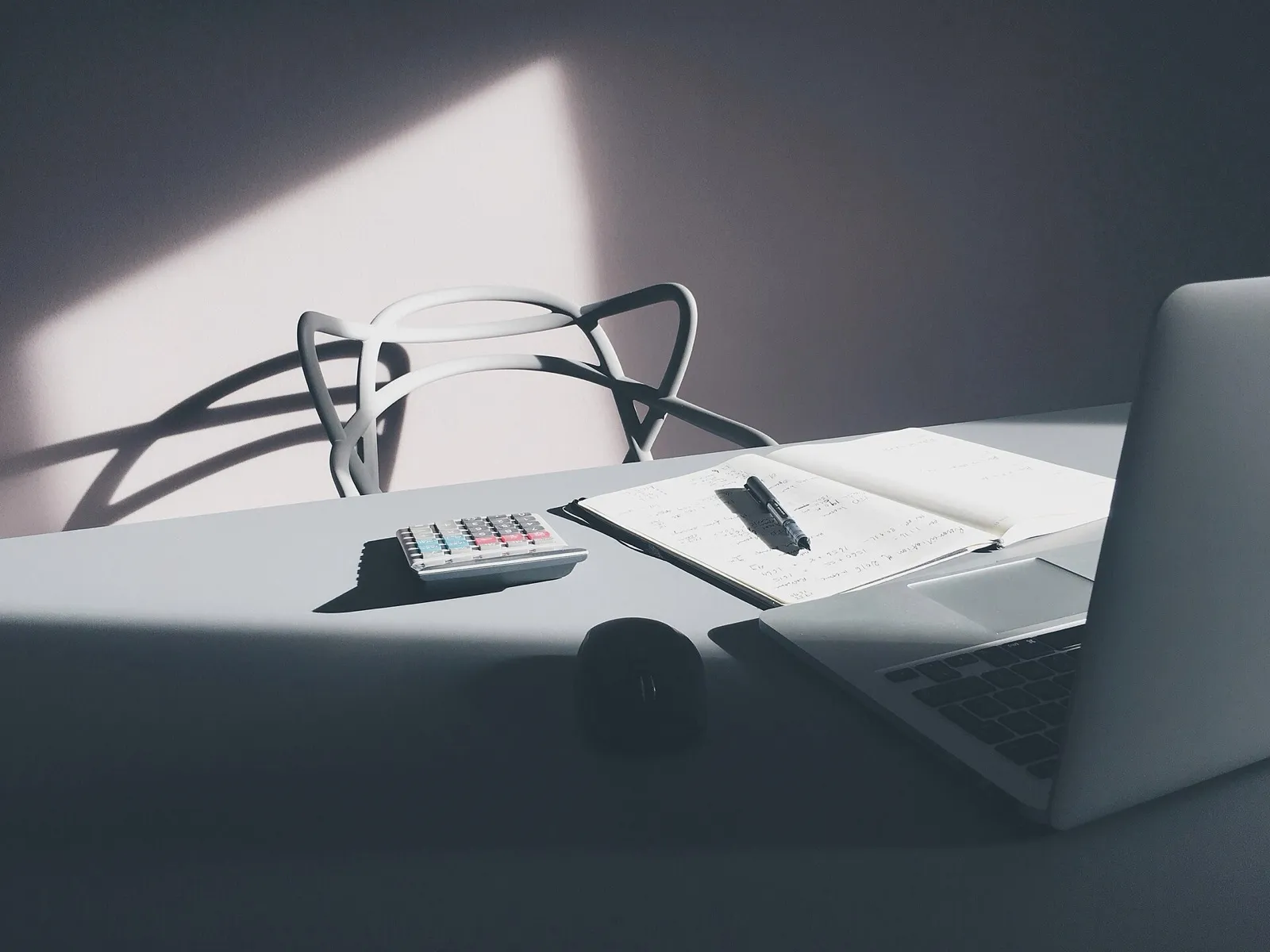 A desk with a calculator, an open notebook of handwritten figures with a pen on it, a mouse, and an open laptop
