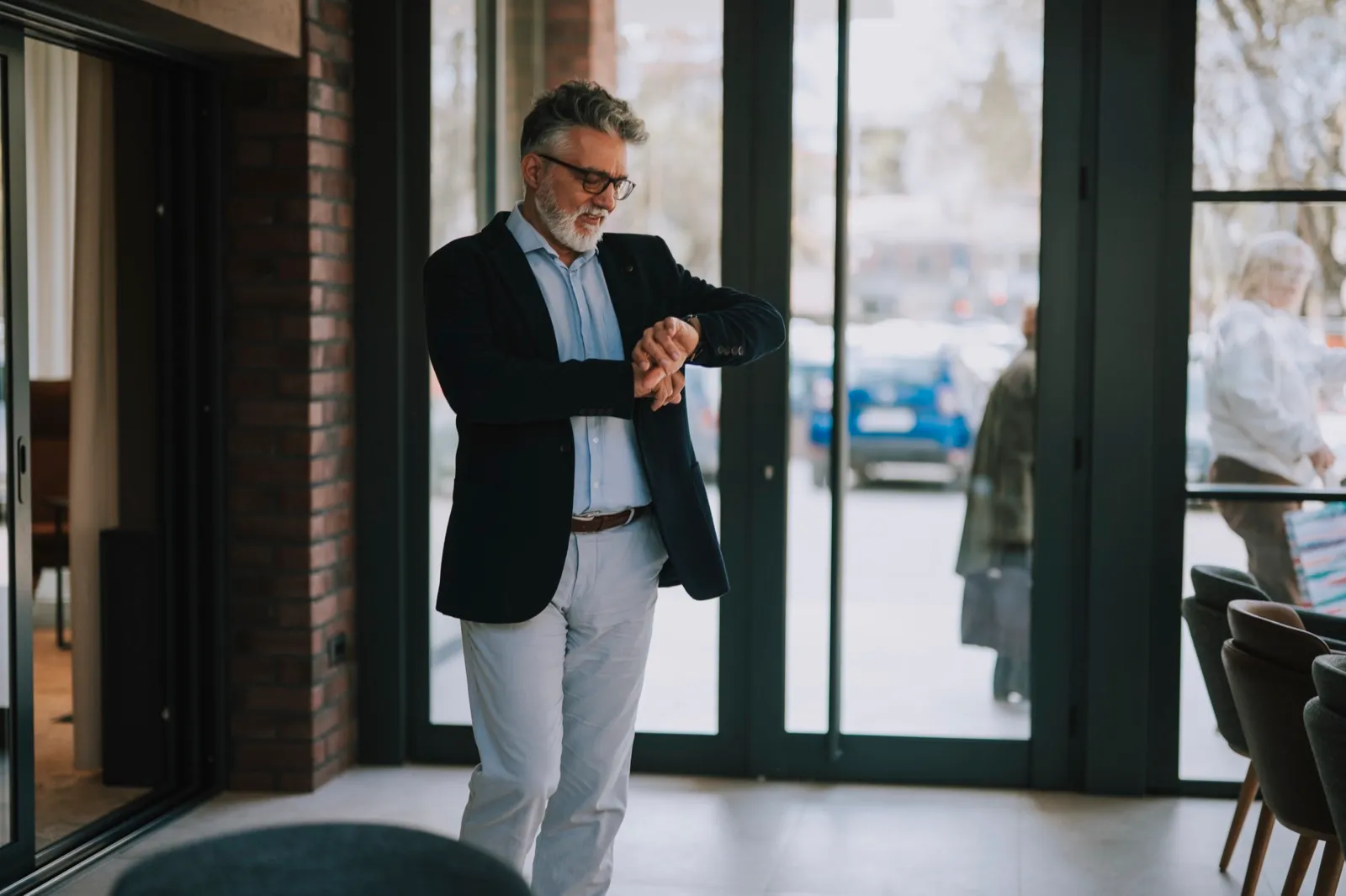 A man in a dark blazer checks his wristwatch while standing by the glass doors of a building lobby