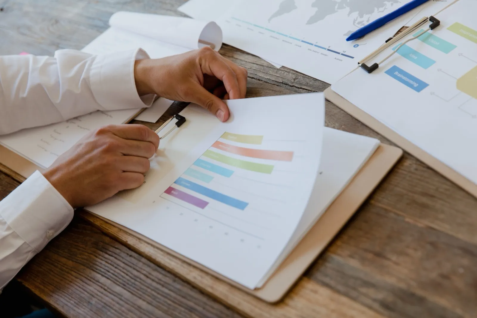 Hands turning the pages of a printed report with a colored bar chart, clipped to a clipboard on a wooden desk