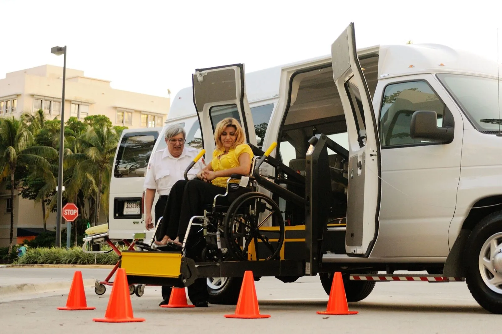 A driver in a white uniform shirt stands beside a woman in a wheelchair on the lift of a white van, with orange cones set out around it