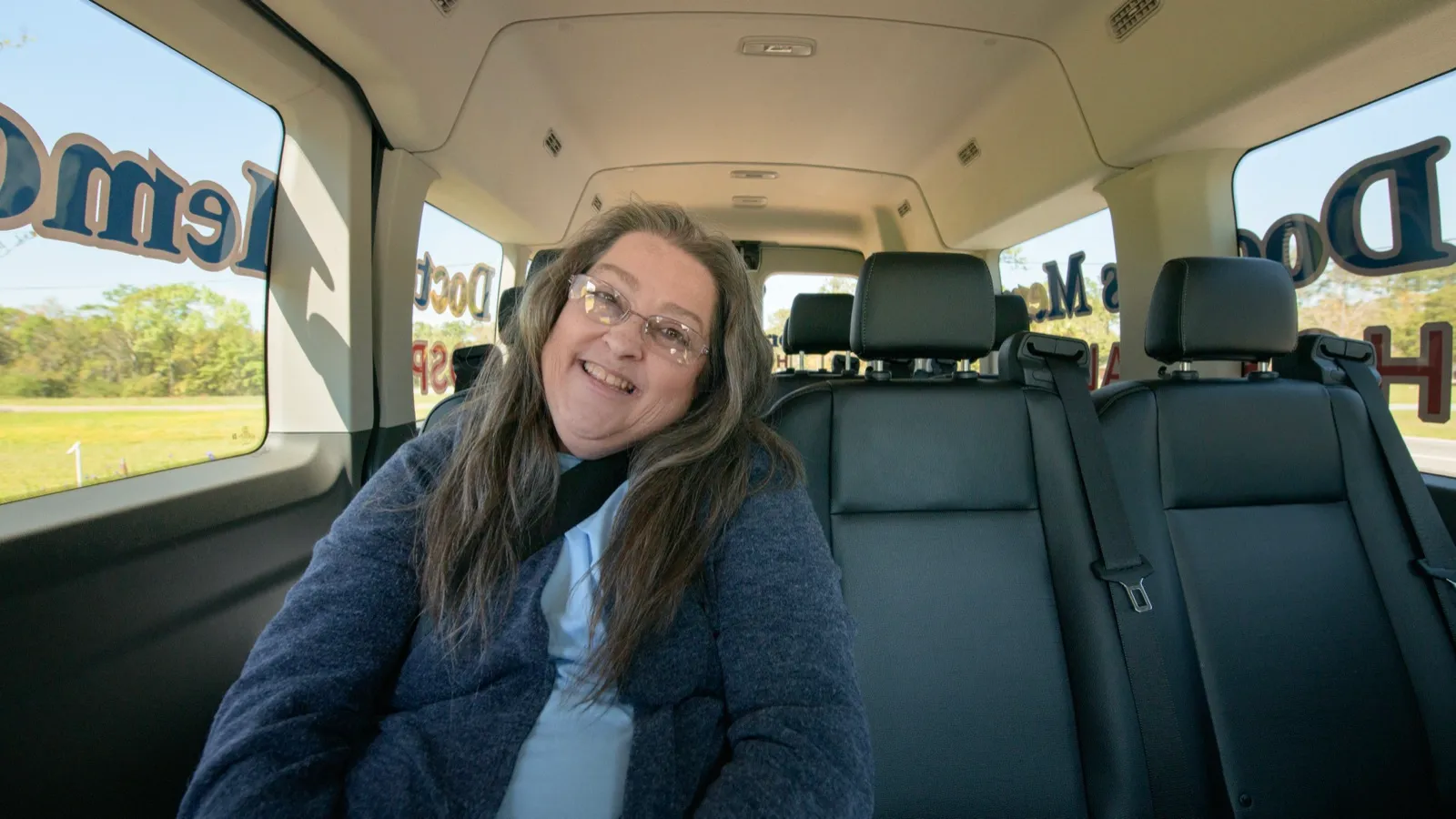 A smiling rider with long gray hair and glasses seated in the back row of a medical transportation van