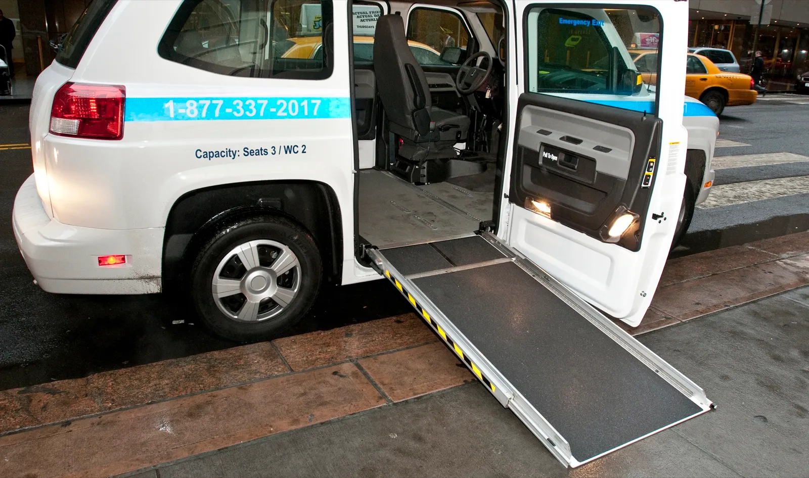 A white wheelchair van parked at a city curb with its side door open and a long ramp folded down to the sidewalk