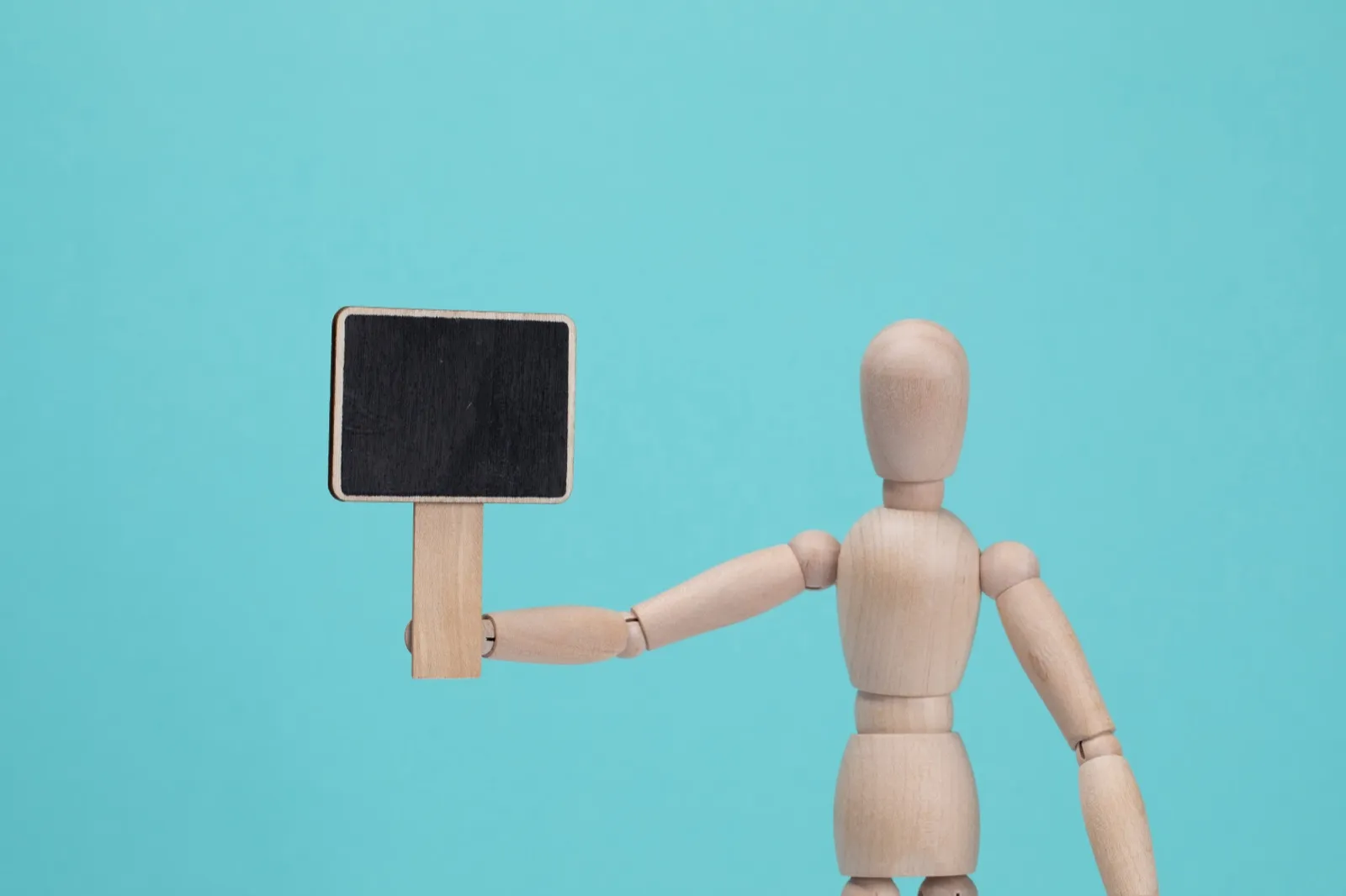 A wooden artist's mannequin holding up a small blank chalkboard sign against a teal background