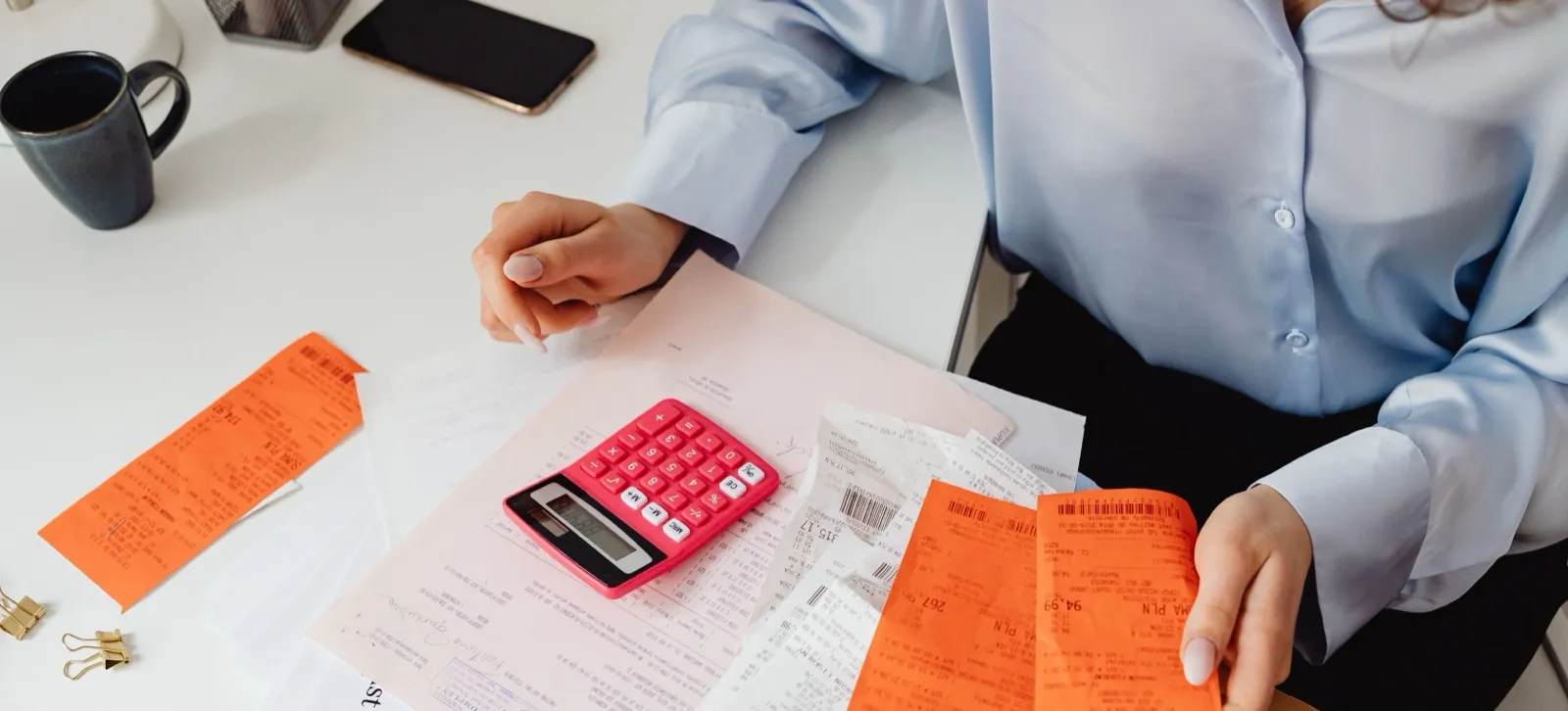 A person at a desk holding orange receipts beside a pink calculator and a spread of printed bills and forms