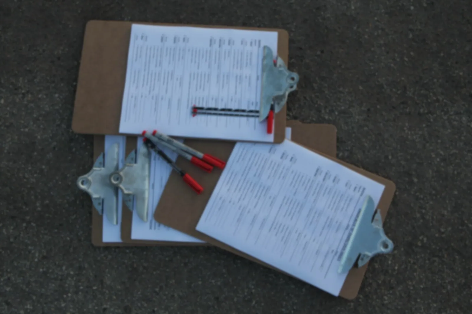 Three clipboards holding printed checklists, with red pens laid across them, on dark pavement