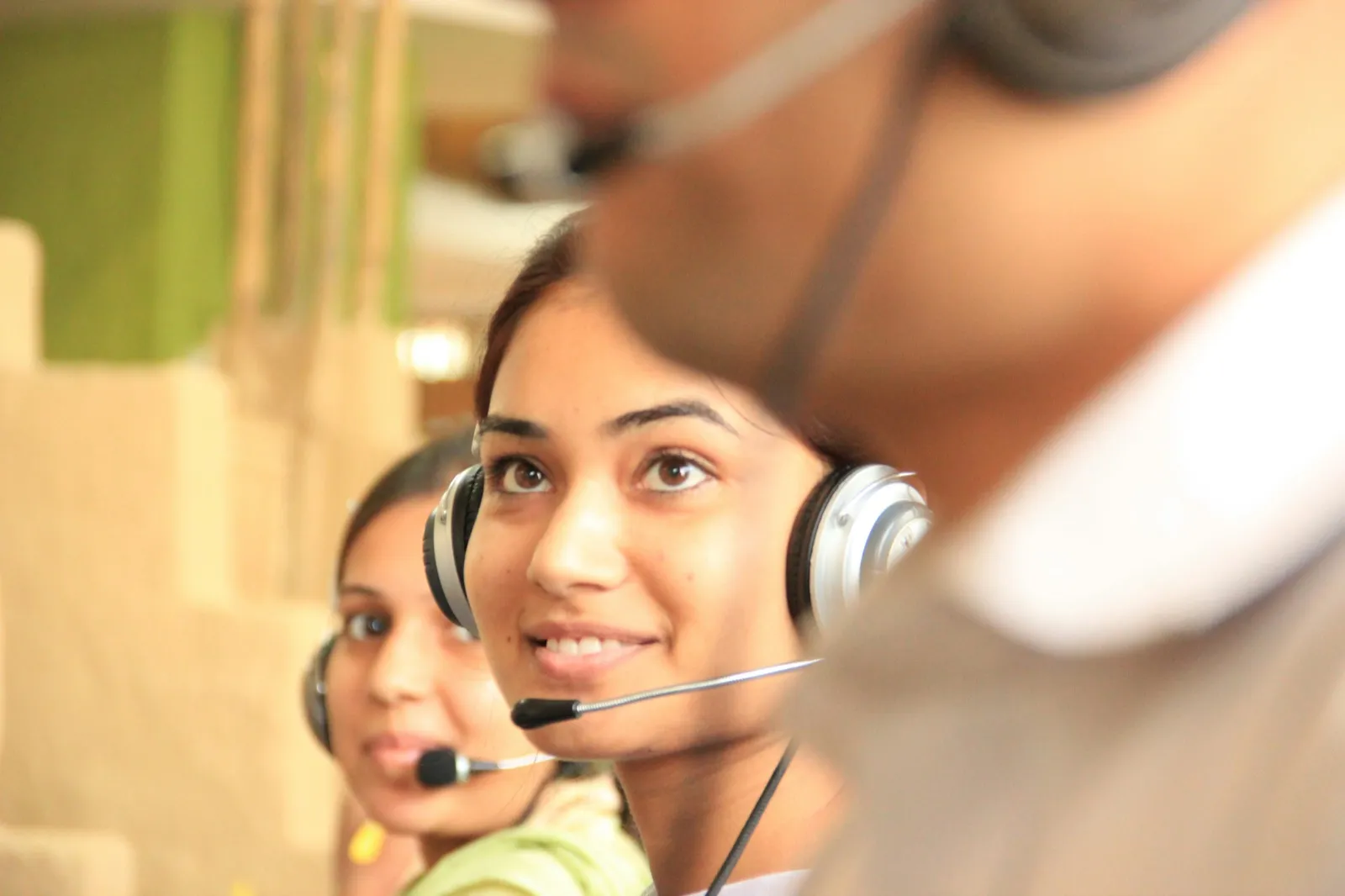 A smiling woman wearing a headset with a microphone, sitting beside a coworker who is also on a headset